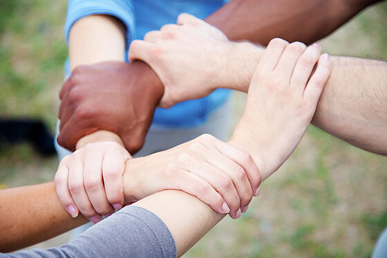 Fünf Erwachsene mit unterschiedlicher Hautfarbe fassen sich gegenseitig am Arm an