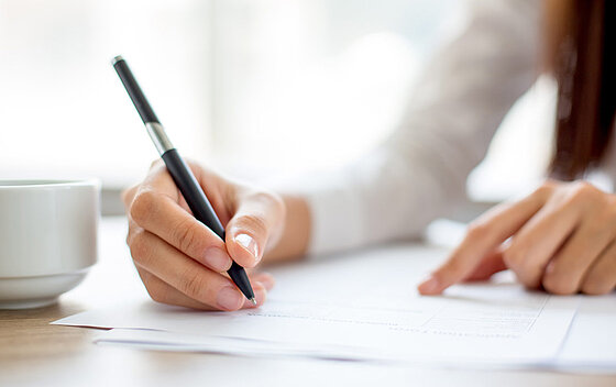 Person schreibt mit Stift etwas auf einem Blatt Papier das auf Tisch liegt