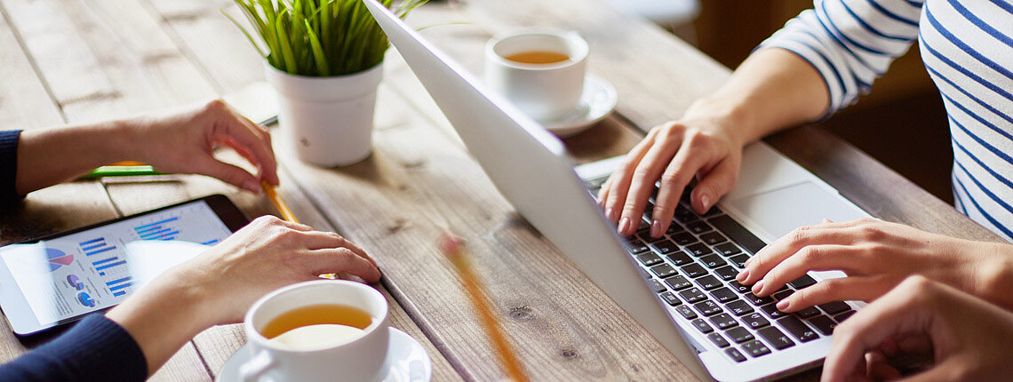 Zwei Personen mit Kaffee am Tisch, eine mit Laptop die andere mit Tablet