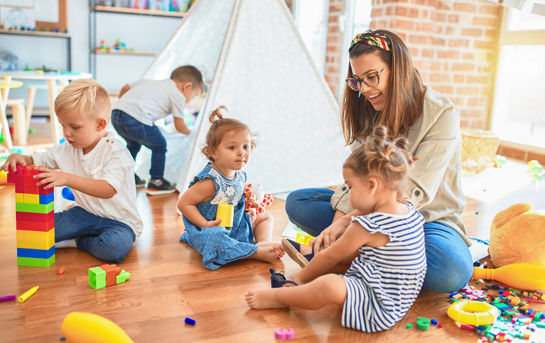 Schöne Lehrerin und Gruppe von Kleinkindern spielen um viele Spielsachen im Kindergarten